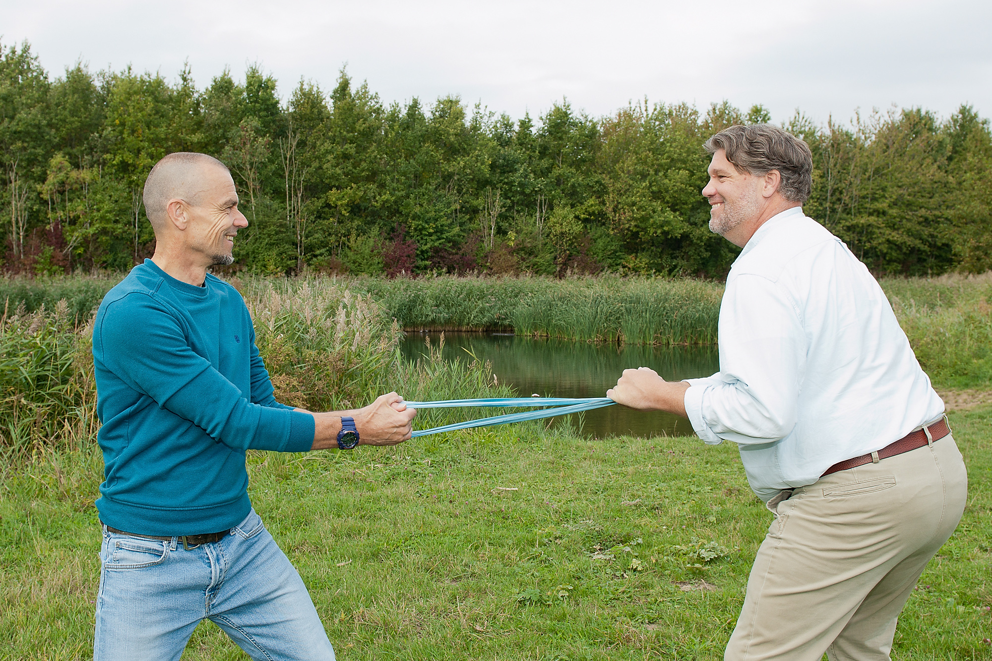 Jan Poul en Erik staan lachend tegenover elkaar en trekken elk aan een lichtblauwe weerstandsband, met gras, water en riet op de achtergrond
