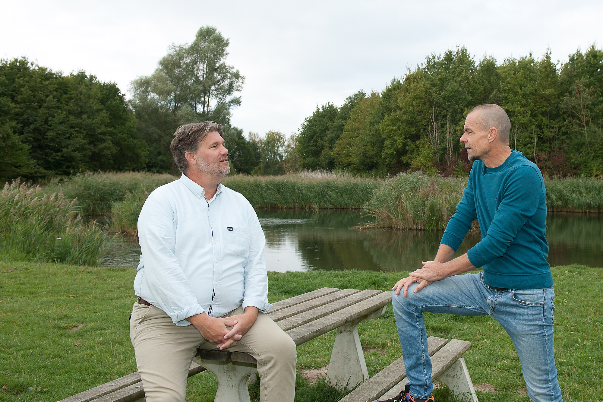 Jan Poul en Erik zitten in gesprek aan een houten tafel op een grasveld met water en riet op de achtergrond