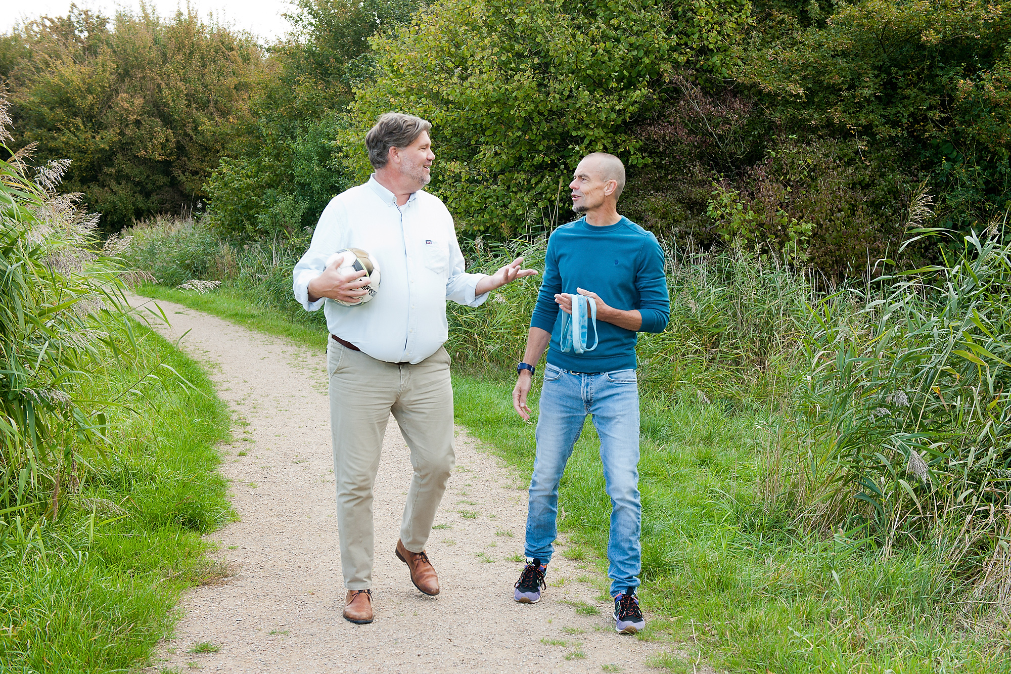 Jan Poul en Erik lopen samen over een zandpad met gras en water op de achtergrond